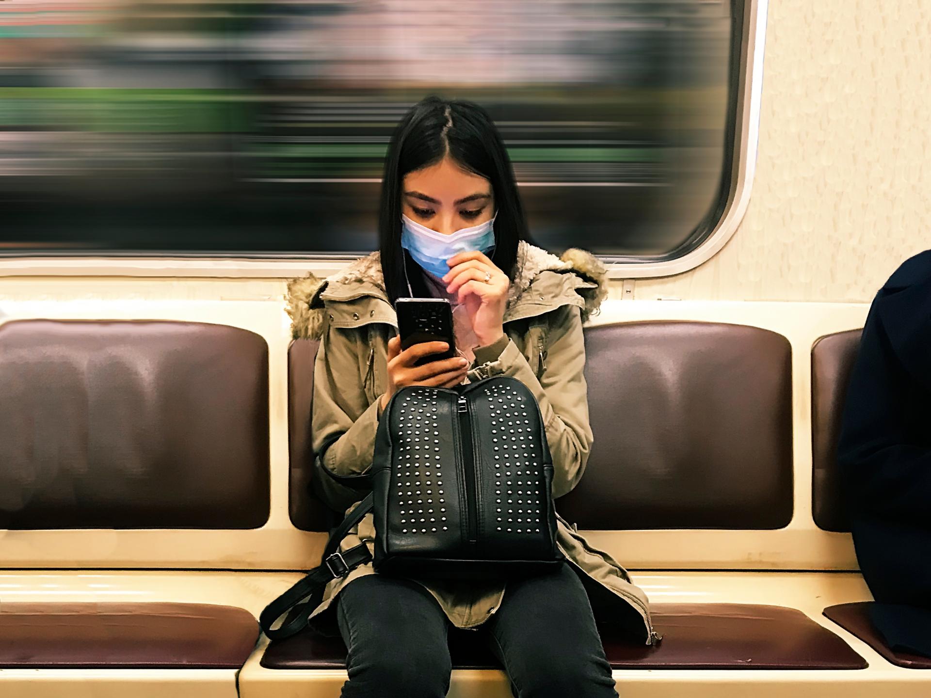 Young woman wearing a medical mask in subway train while texting on mobile phone