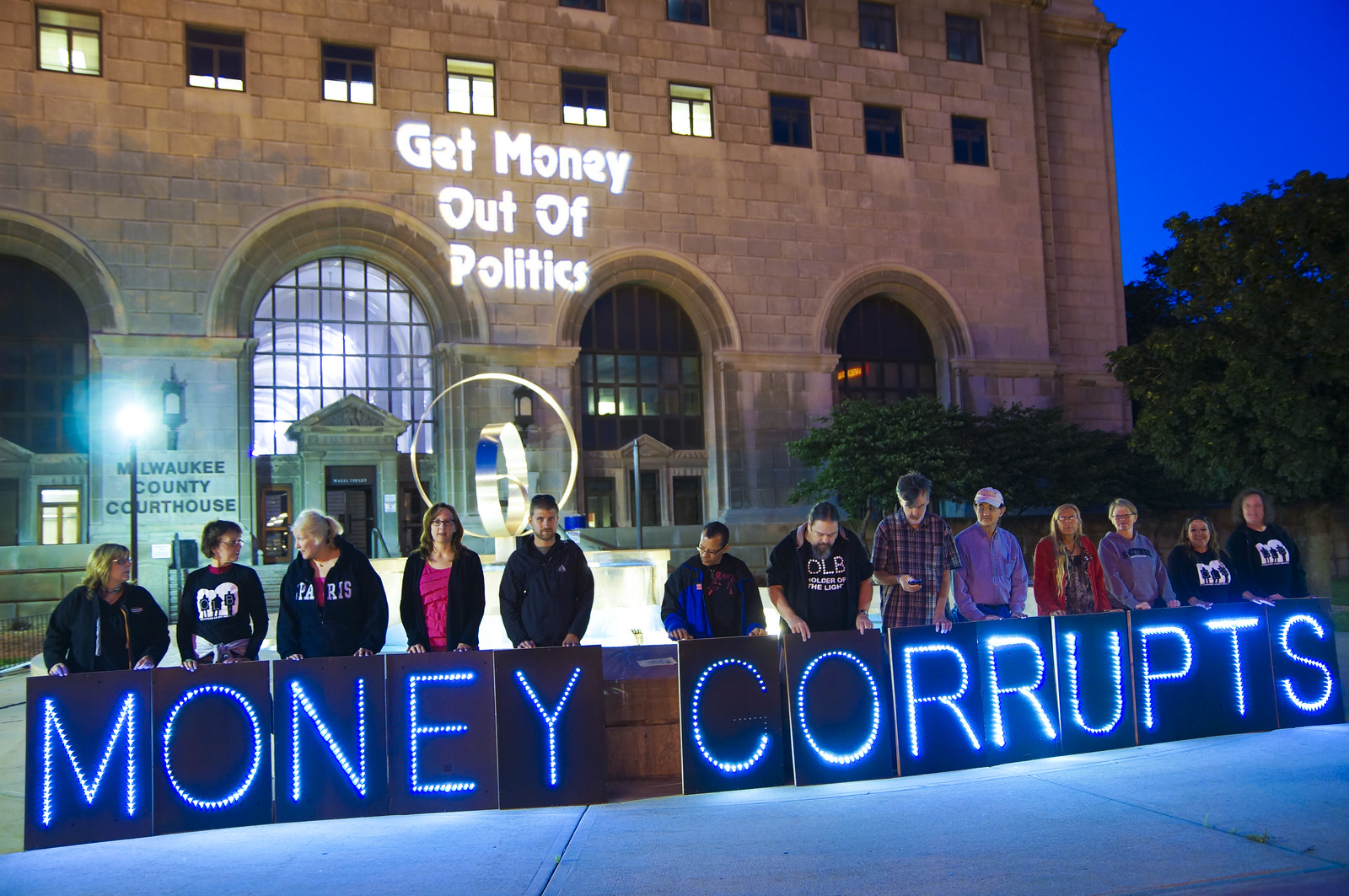People holding a sign that read, 'Money Corrupts' outside of Milwaukee County Court. 'Get money out of politics' is projected on the wall behind them.
