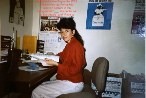 Deborah Levine Roy at the IFES office in Port-au-Prince with election posters in the background: "____ sits on the will of the people" and "Right to Vote"