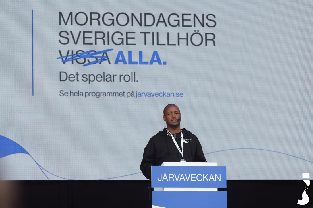 Ahmed Abdirahman speaking on the main stage at Järvaveckan 2024. The backdrop behind him reads: ‘The Sweden of tomorrow belongs to all’ — with the word ‘some’ visibly crossed out. Photo by:&nbsp;Järvaveckan/Avandimedia (2024)